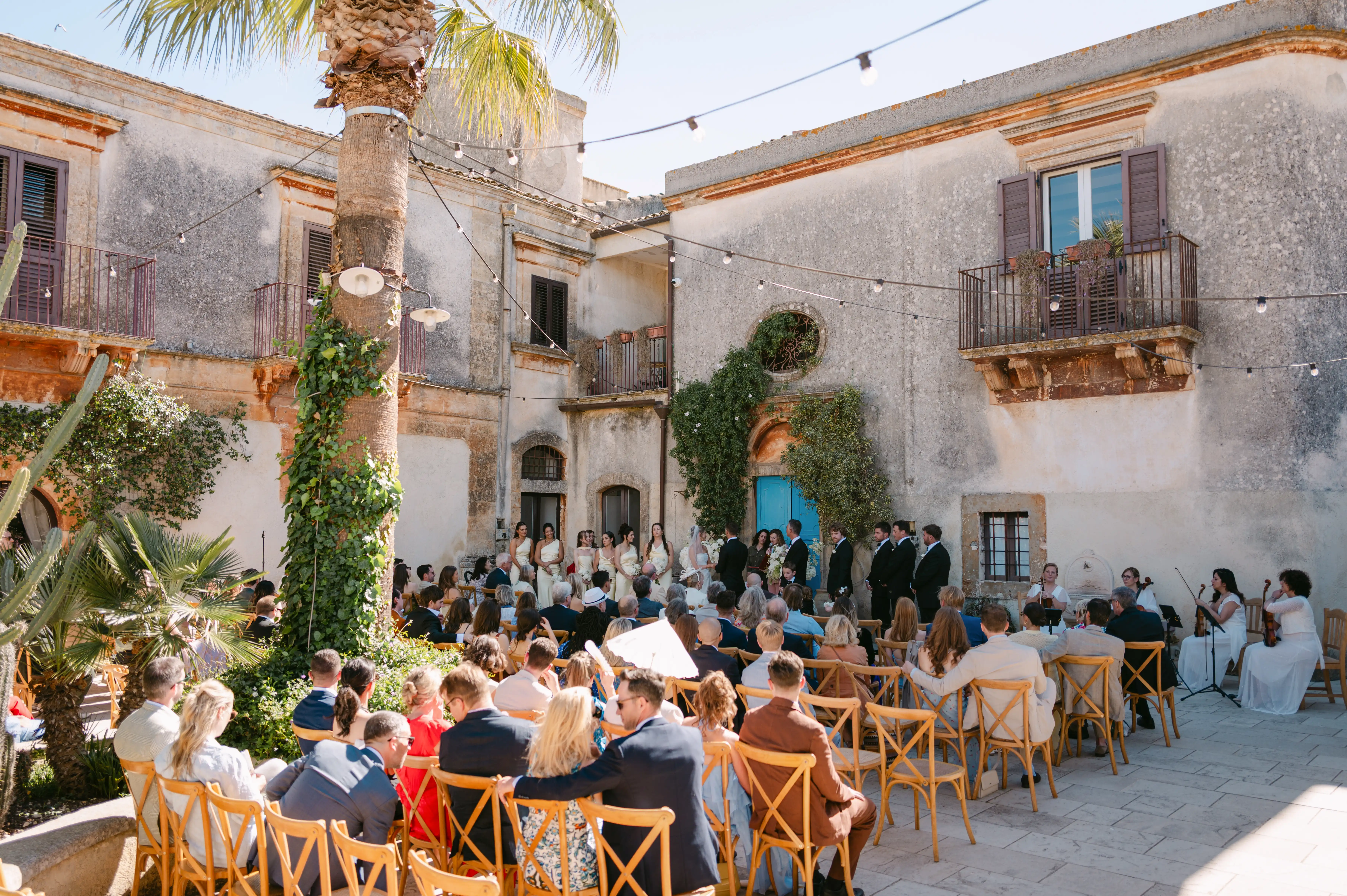 Luxury outdoor wedding ceremony in a historic Sicilian courtyard with guests seated under palm trees and string lights.